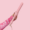 A pink curling wand with a medium barrel, held in a hand against a pink background.