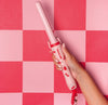 A hand holding a pink curling wand with cherry design against a pink and red checkered background.
