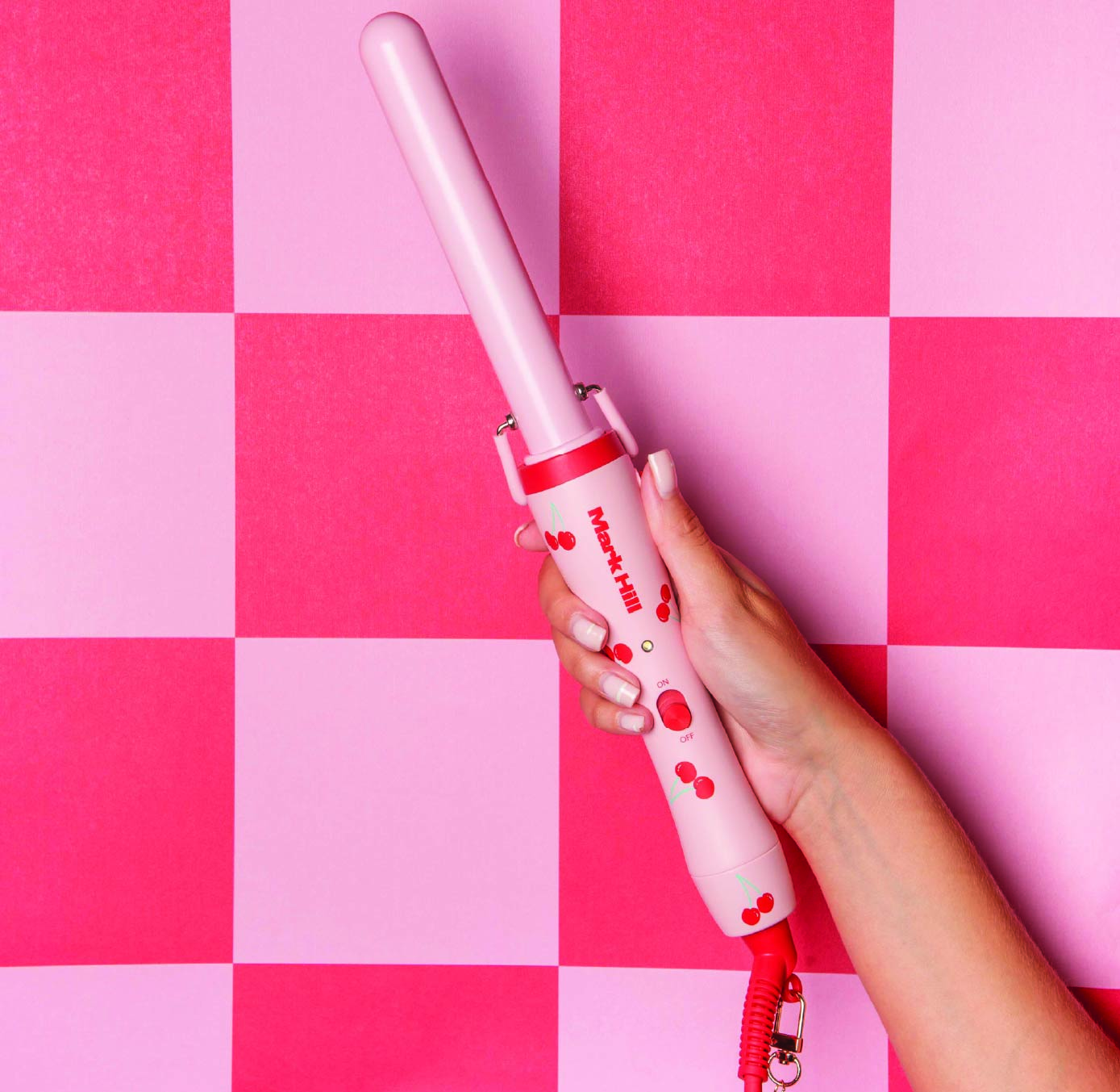 A hand holding a pink curling wand with cherry design against a pink and red checkered background.