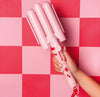 A hand holding a pink curling wand with a cherry design on a red and pink checkered background.