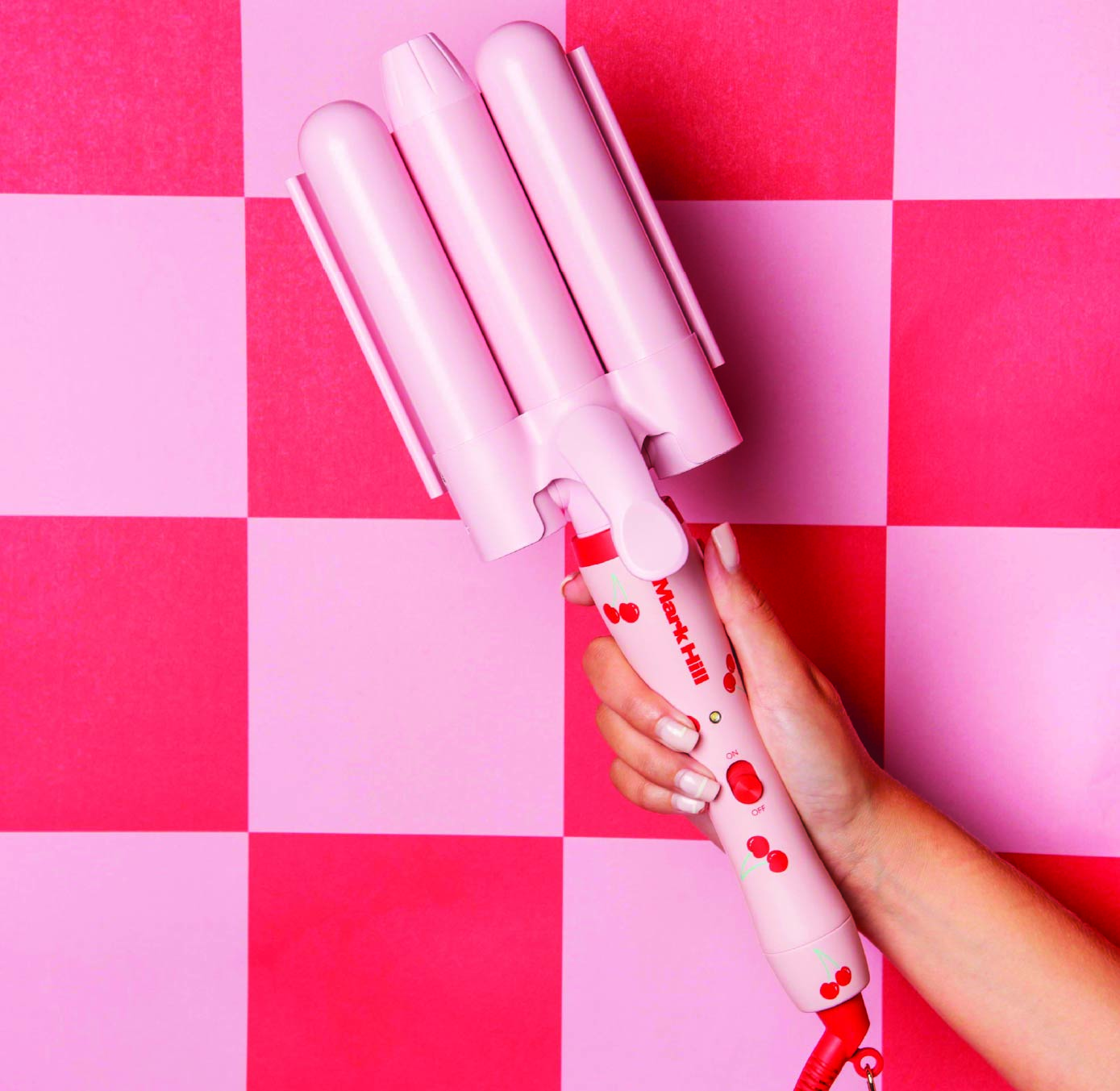 A hand holding a pink curling wand with a cherry design on a red and pink checkered background.